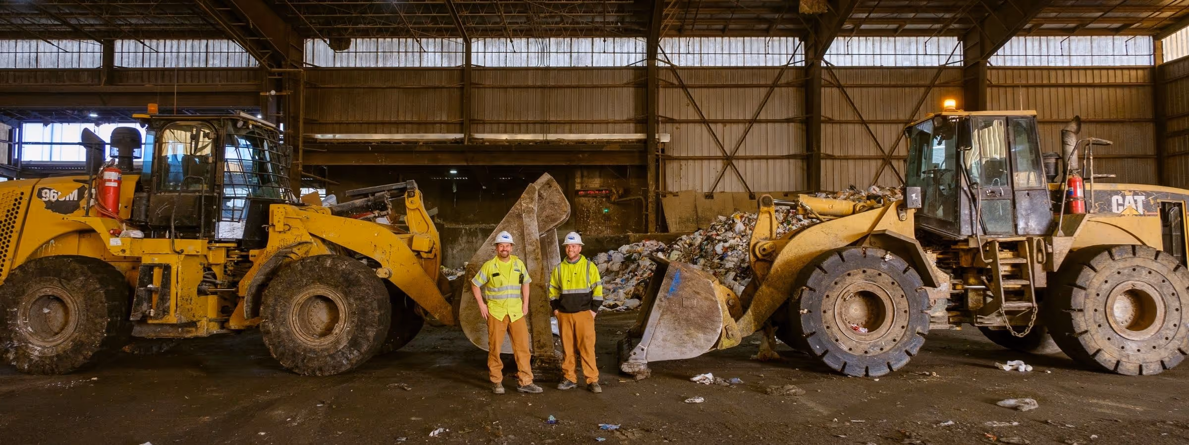 Two workers in high-visibility gear stand between large yellow wheel loaders in a recycling facility.