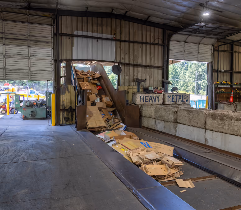 Cardboard boxes on conveyor belt inside recycling facility with 'HEAVY METALS' sign in background.