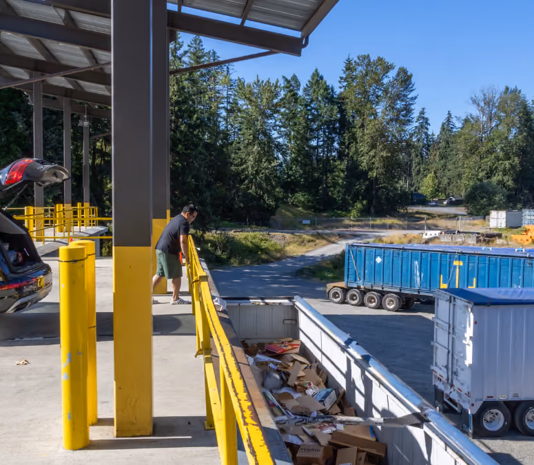 Man wearing a mask stands on a yellow-railed platform next to an open car trunk at a transfer station.