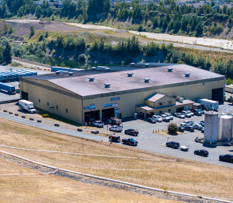 Aerial view of a large industrial building with multiple vehicles parked outside and wooded areas in the background.