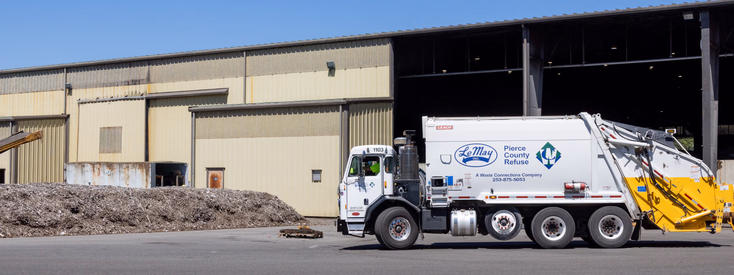 LeMay Pierce County Refuse garbage truck parked outside a large industrial building near a pile of debris.