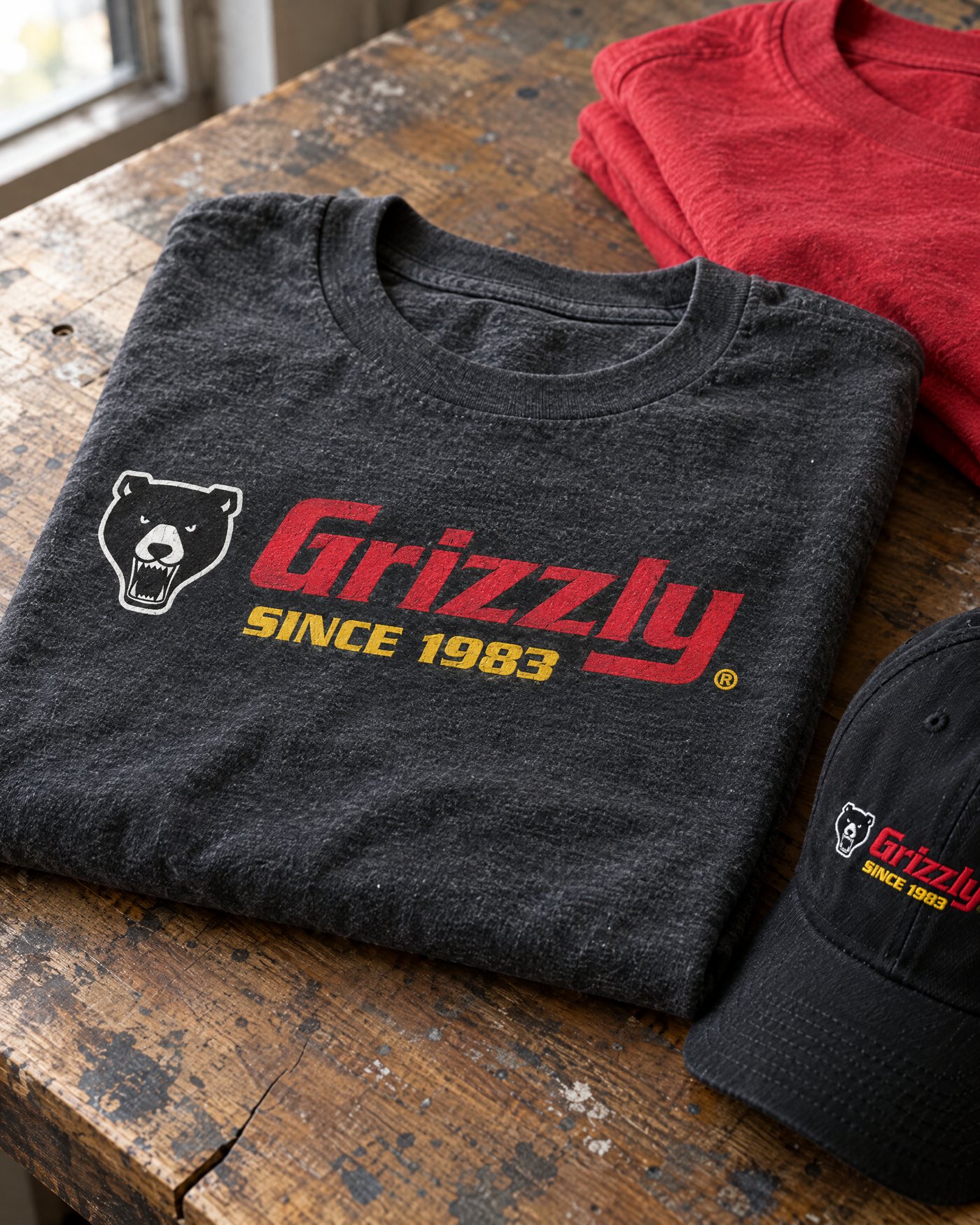 Grizzly heritage-color tee in charcoal with red and gold print, next to a red tee and black cap