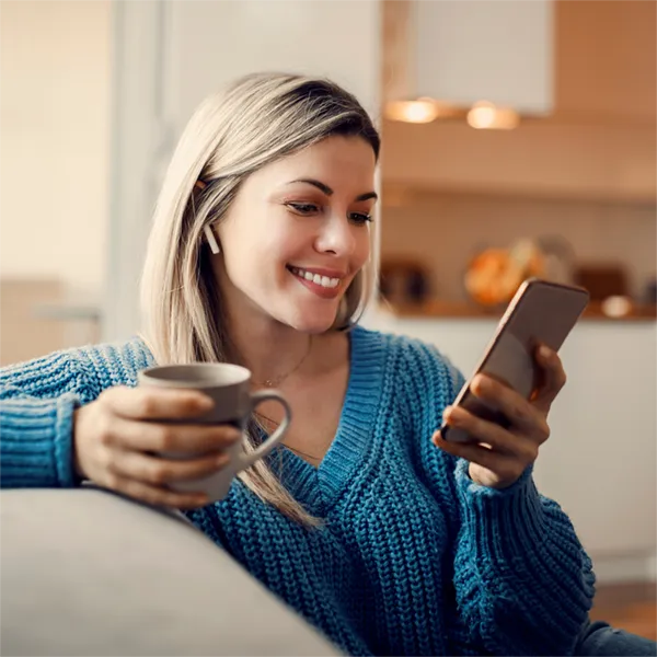Smiling woman in blue sweater holding a cup and looking at her smartphone indoors.