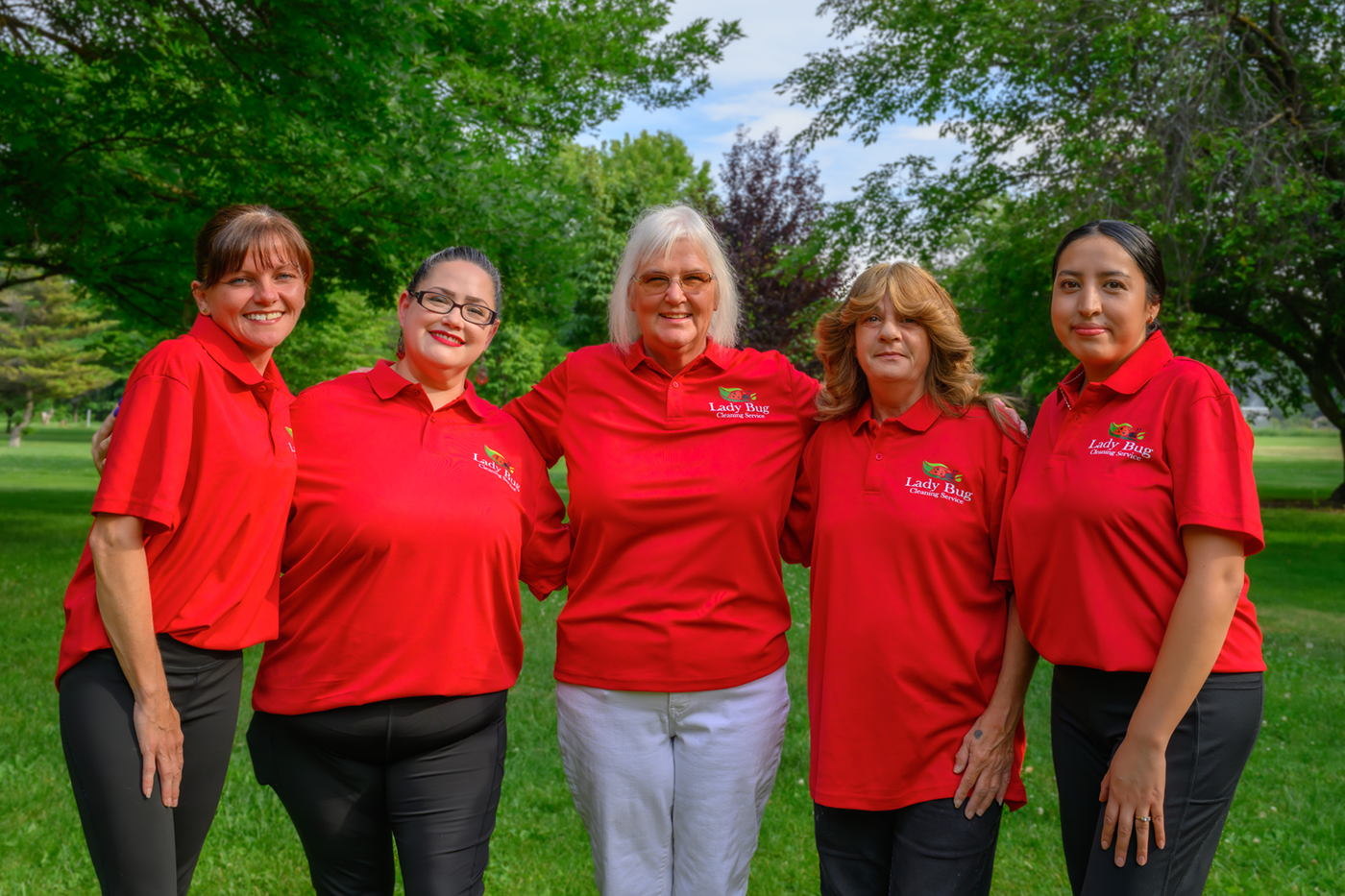 Five women in red Lady Bug Cleaning Service shirts standing outdoors with trees in the background.