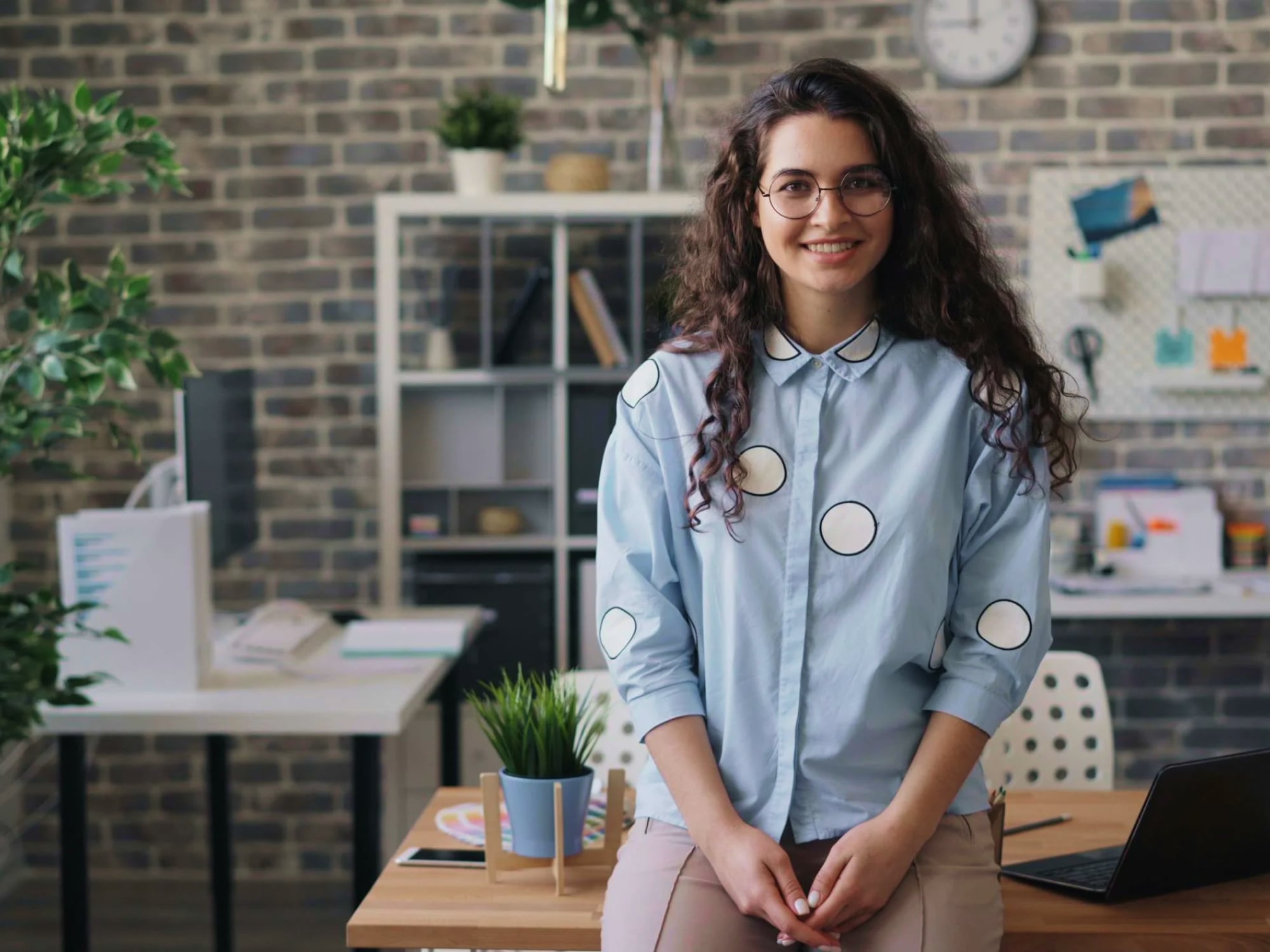 Smiling woman with curly hair and glasses in a polka dot shirt sitting on a desk in a modern office.