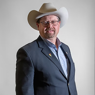 Man in glasses wearing a light cowboy hat, dark blazer, and blue shirt against a plain background.