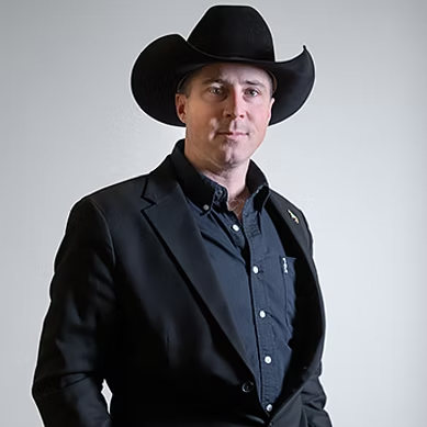 Man wearing a black cowboy hat and suit jacket against a plain gray background.