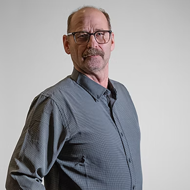 Middle-aged man with glasses and mustache wearing a gray button-up shirt against a plain background.