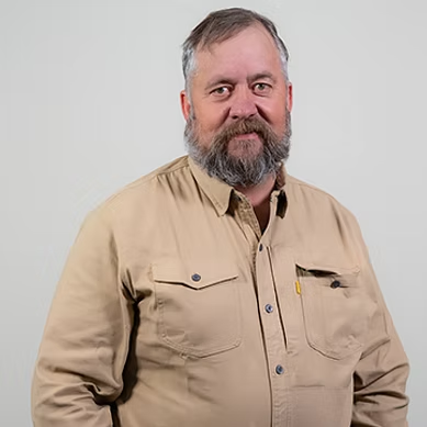 Middle-aged man with gray beard wearing a tan button-up shirt against a light gray background.