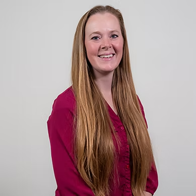 Smiling woman with long straight hair wearing a magenta blouse against a light gray background.