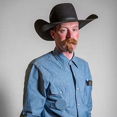 Man with a mustache and goatee wearing a black cowboy hat and blue button-up shirt.