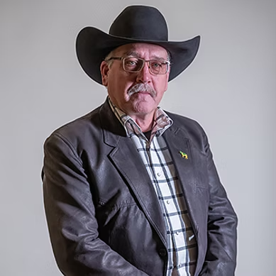 Older man wearing glasses, a black cowboy hat, plaid shirt, and dark leather jacket against a gray background.
