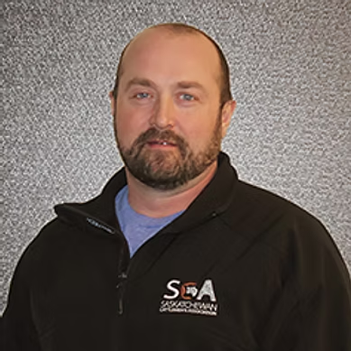 Bearded man wearing a black jacket with an embroidered logo on gray textured background.