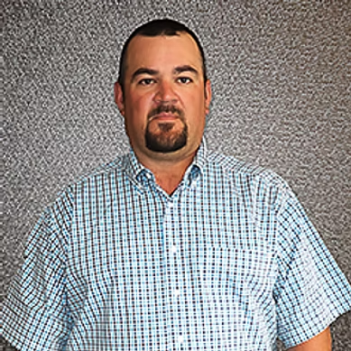 Portrait of a man with short dark hair and goatee wearing a blue checkered shirt against a textured wall.