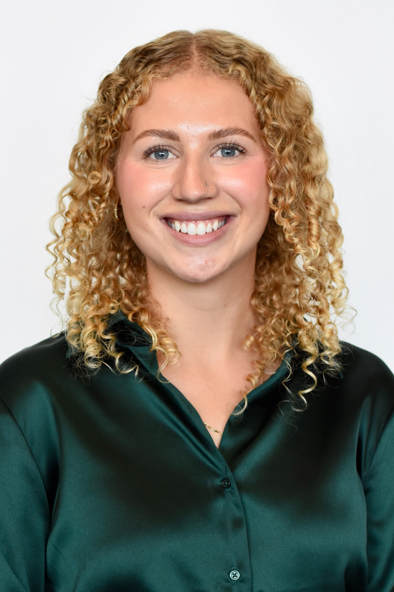 Smiling woman with curly blonde hair wearing a dark green satin blouse on a white background.