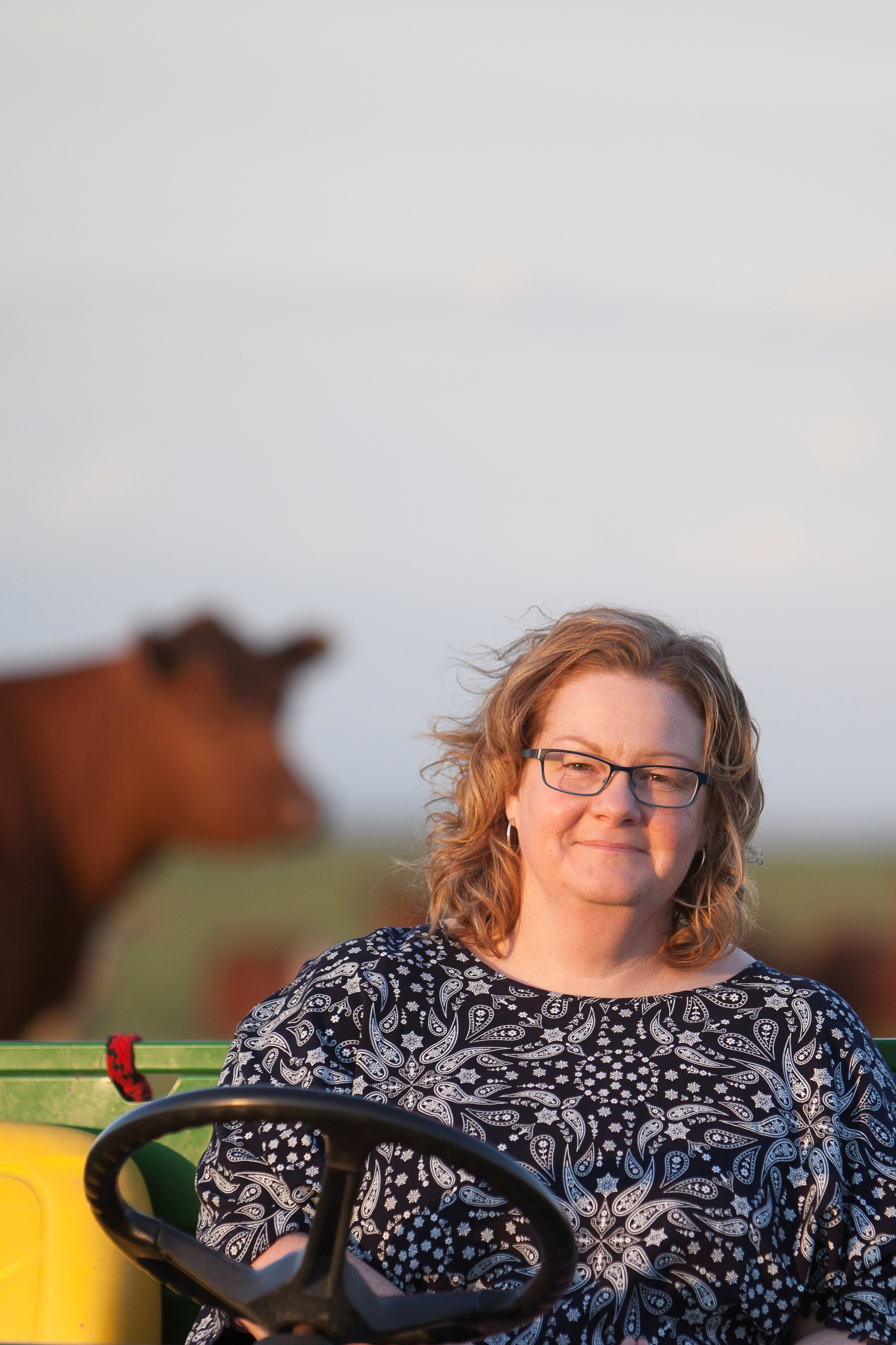 Woman wearing glasses sitting on a tractor with a cow in the blurred background.