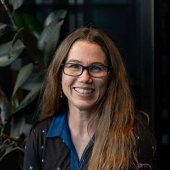 Woman with long brown hair wearing glasses and a blue collared shirt smiling against a dark background with green plants.