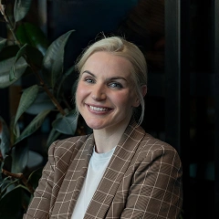 Smiling woman with blonde hair tied back, wearing a brown checkered blazer and white shirt, sitting indoors with green plants in the background.