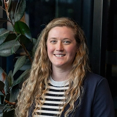 Smiling woman with long wavy blonde hair, wearing a striped shirt and dark blazer, standing indoors near leafy plants and a dark window.