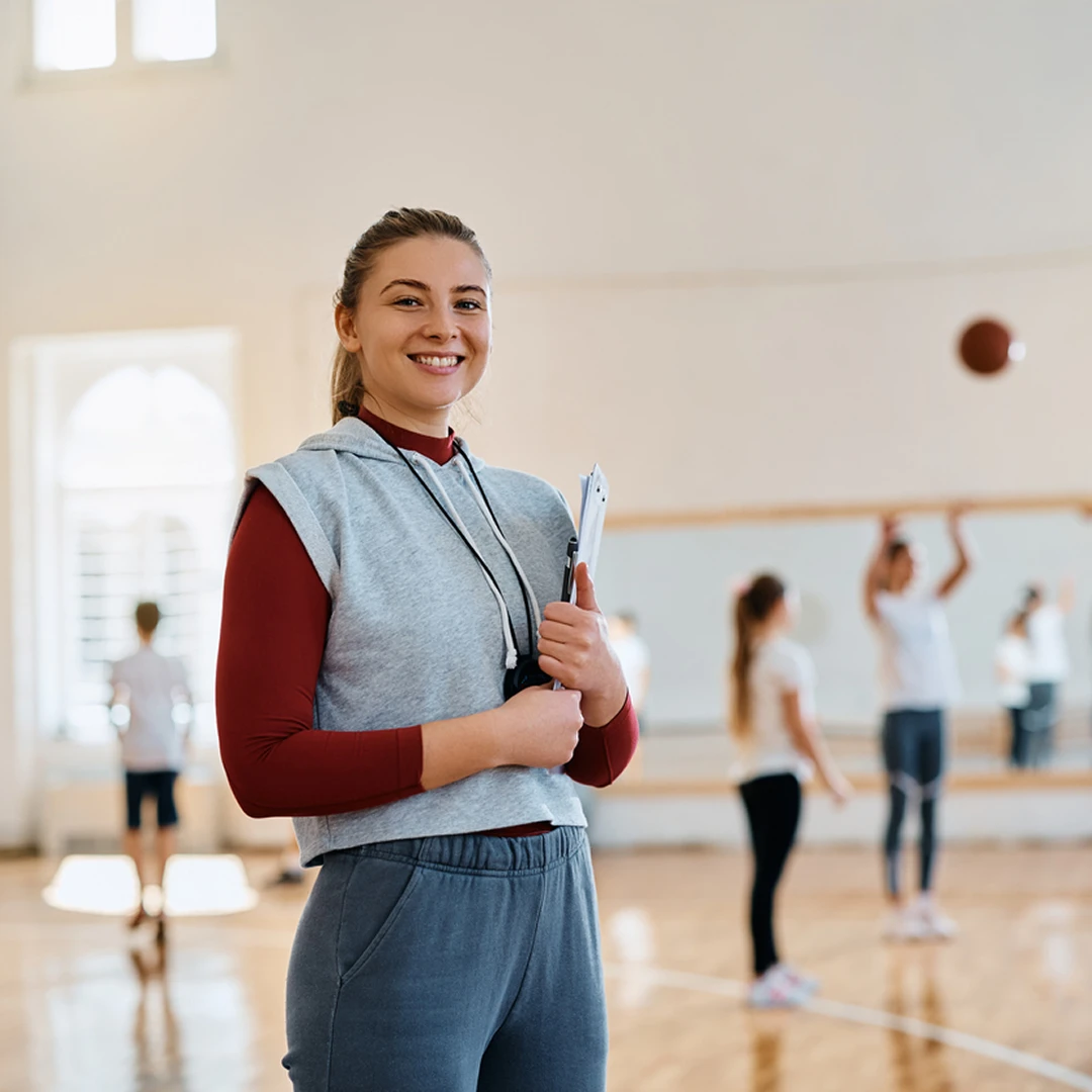 Female coach standing in a gymnasium holding a clipboard and whistle around her neck, smiling at the camera, with children playing basketball in the background.