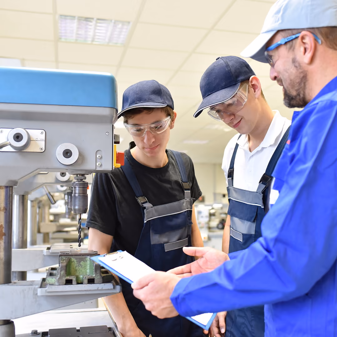 Two young men wearing safety glasses, caps, and work overalls look at a clipboard held by an instructor in a blue uniform near a large machine drill press in a workshop.