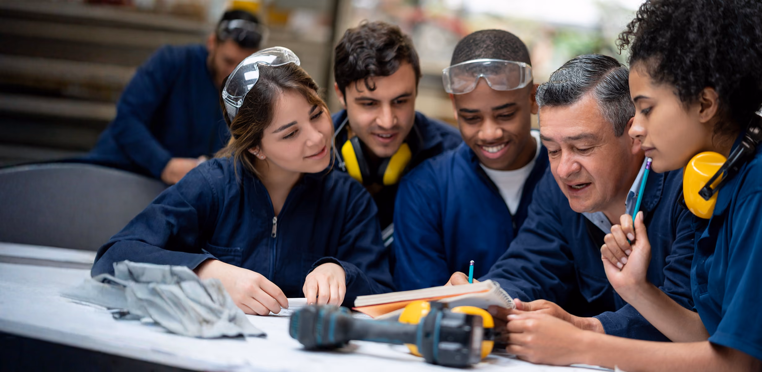 A group of four diverse students in blue work uniforms gathered around a table with a male instructor, all looking at notes and tools while engaged in a hands-on learning activity.