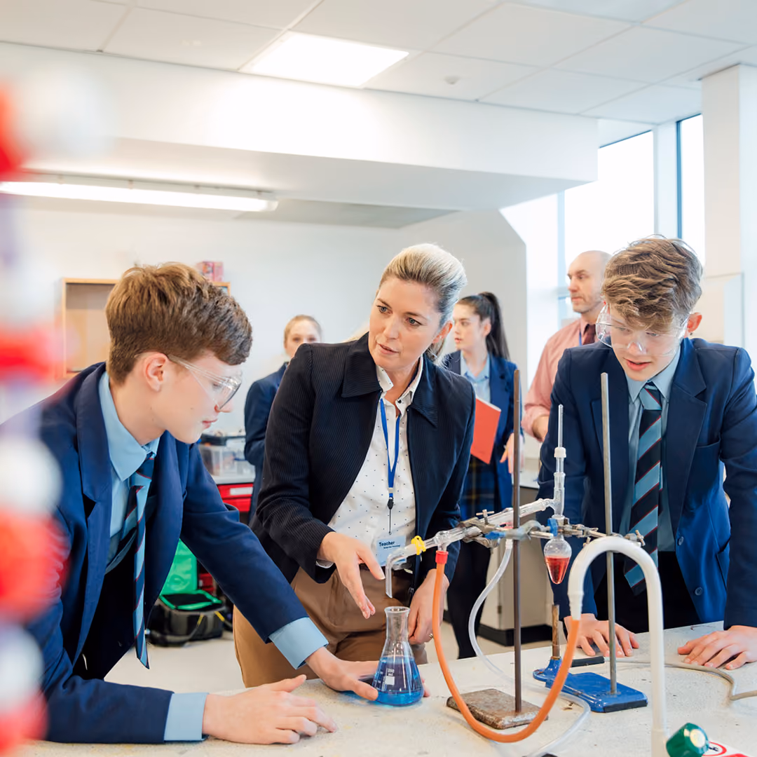 Female teacher instructing two male high school students in a science lab with chemical glassware and blue liquid during an experiment, all wearing school uniforms and safety glasses.