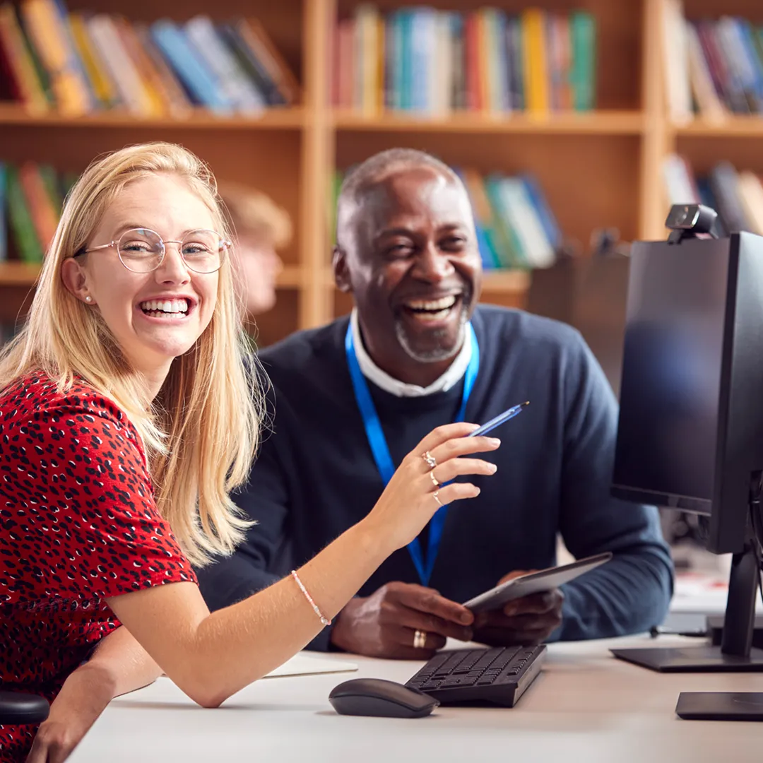 Two coworkers, a woman with glasses in a red patterned shirt and a man in a dark sweater with a blue lanyard, smiling and working together at a desk with a computer, keyboard, and tablet in a library setting with bookshelves in the background.
