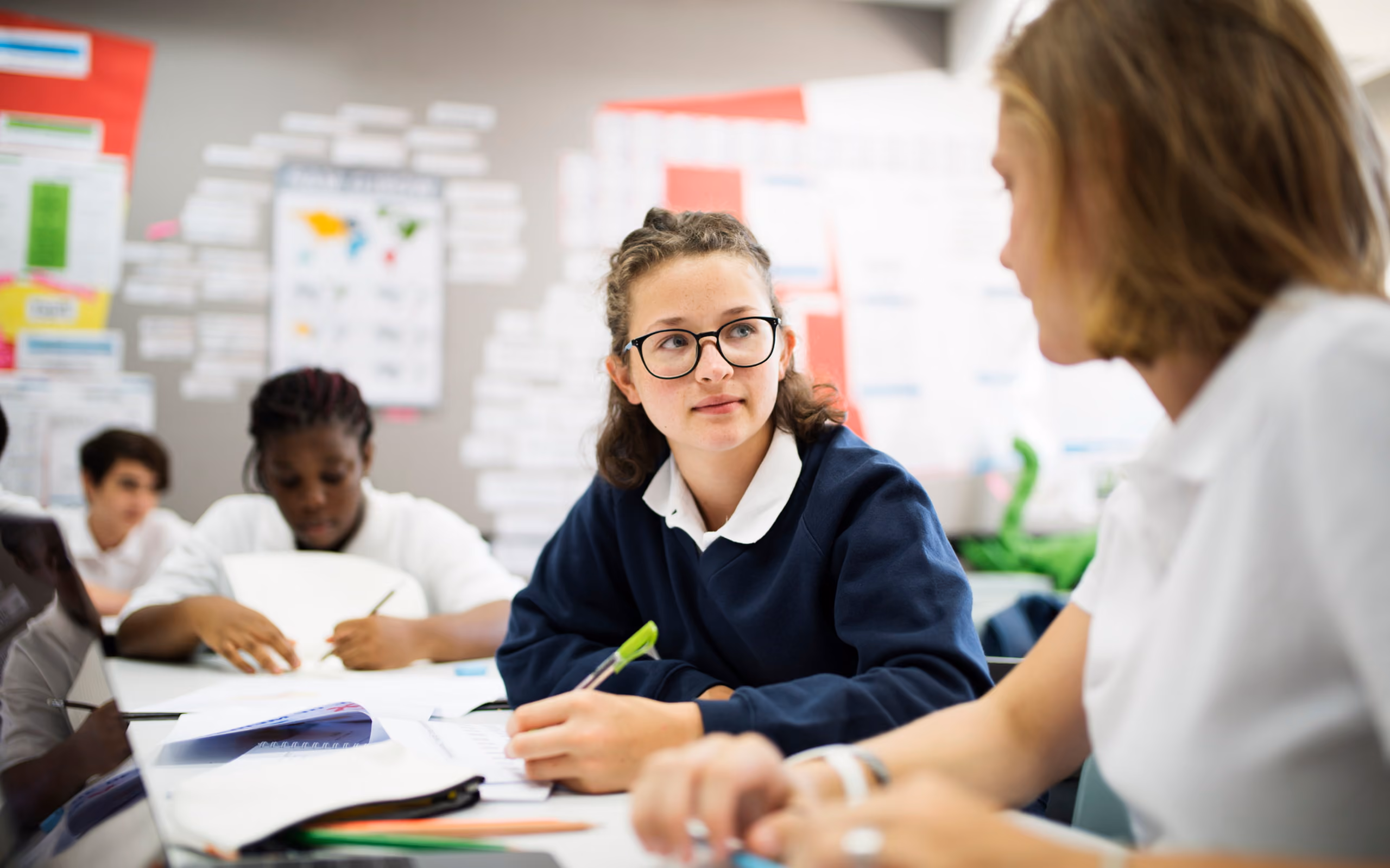 Female student wearing glasses and a navy sweater attentively listening to a teacher in a classroom setting with other students working in the background.