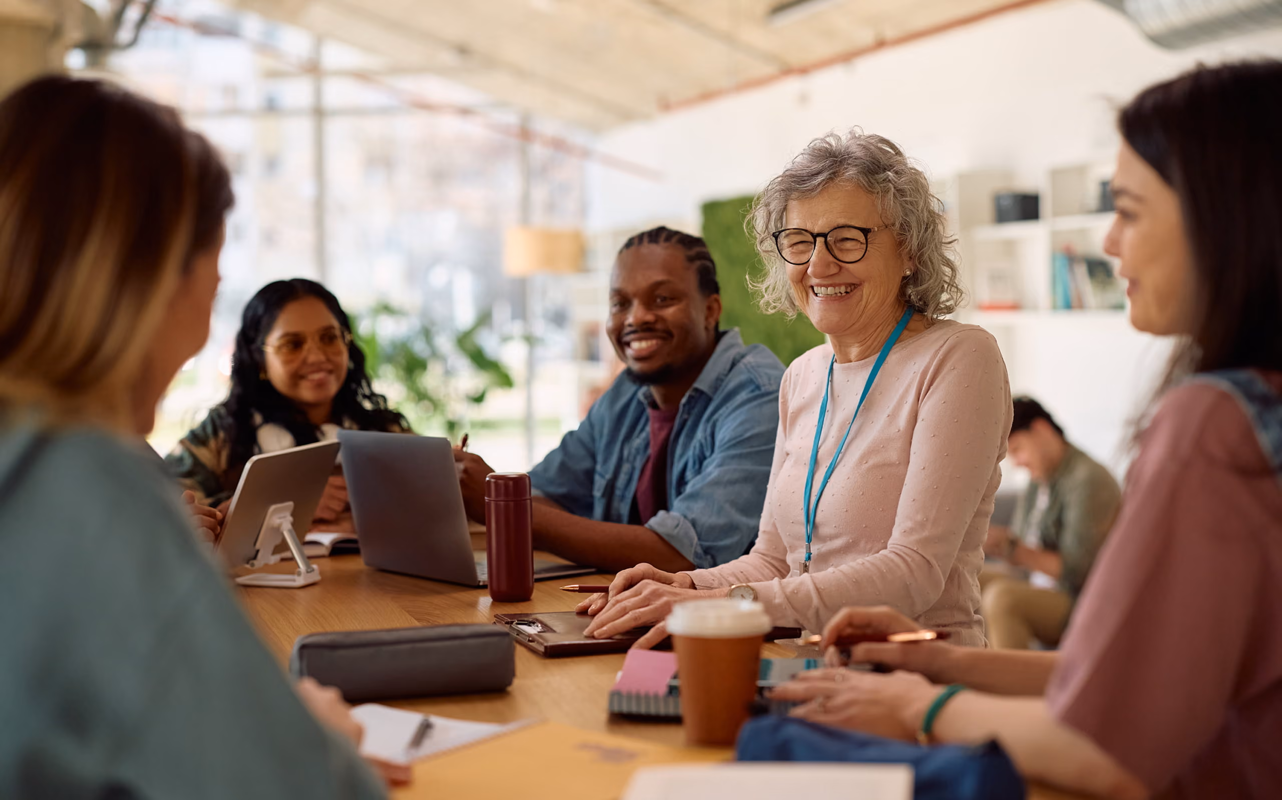 A diverse group of five people sitting around a table in a bright room, smiling and engaging in discussion with laptops, notebooks, and coffee cups.