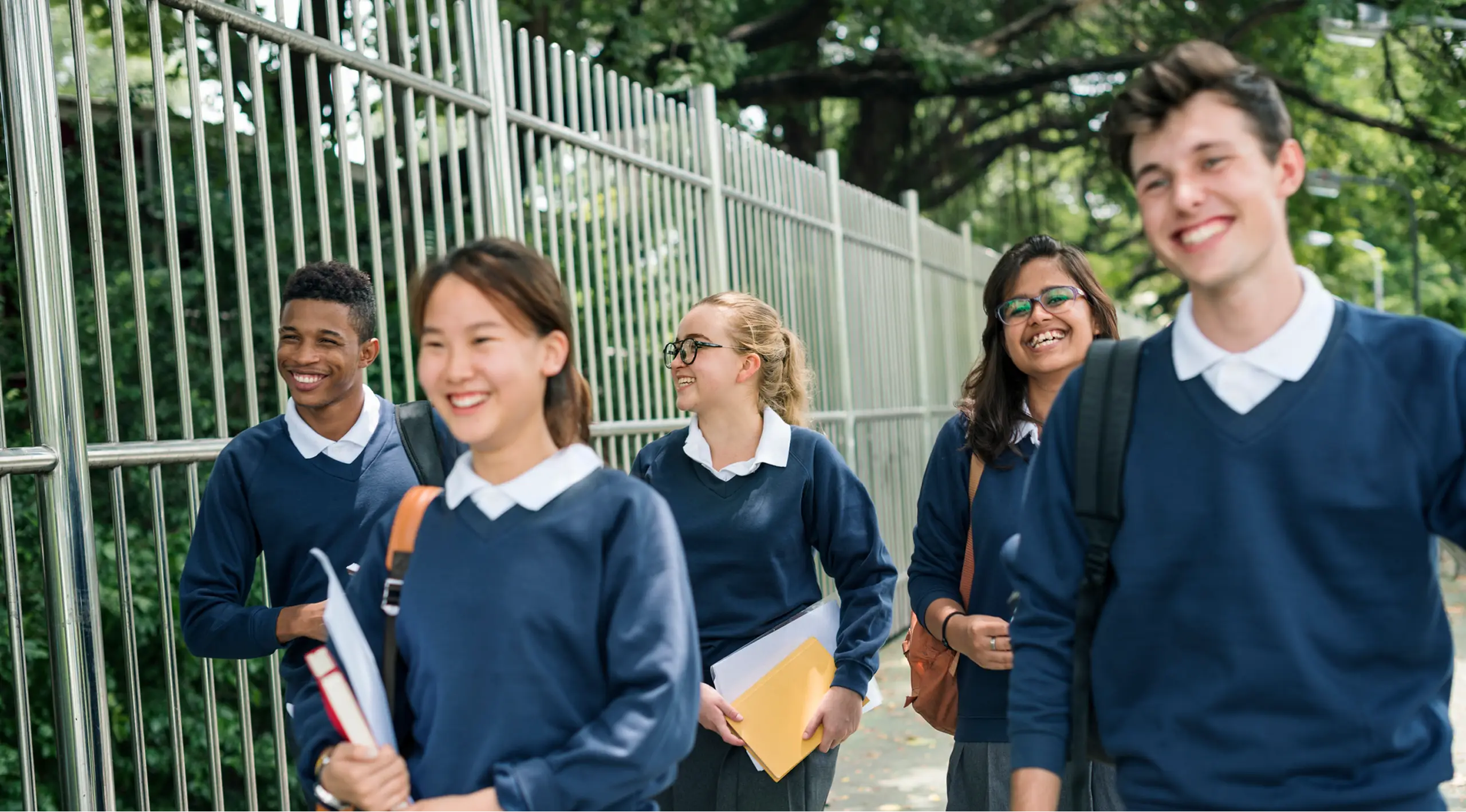 Group of five smiling high school students wearing navy blue sweaters and white collared shirts walking outside near a metal fence with trees in the background, carrying books and backpacks.