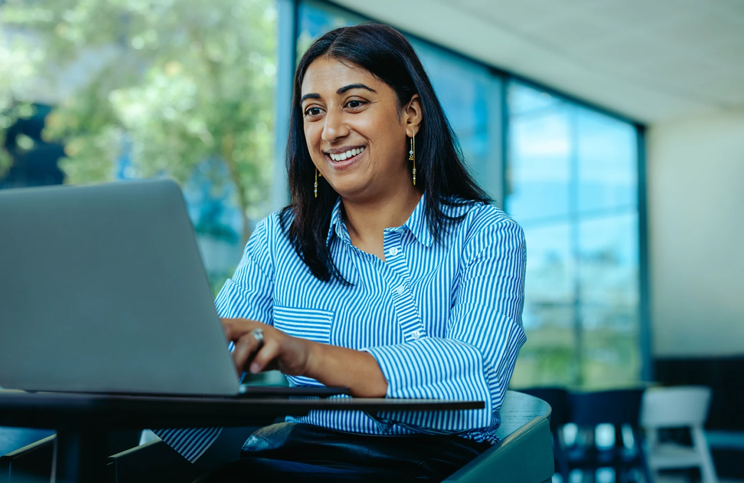 Smiling woman with long dark hair wearing a blue and white striped shirt working on a laptop at a table in a bright modern indoor space with large windows and greenery outside.