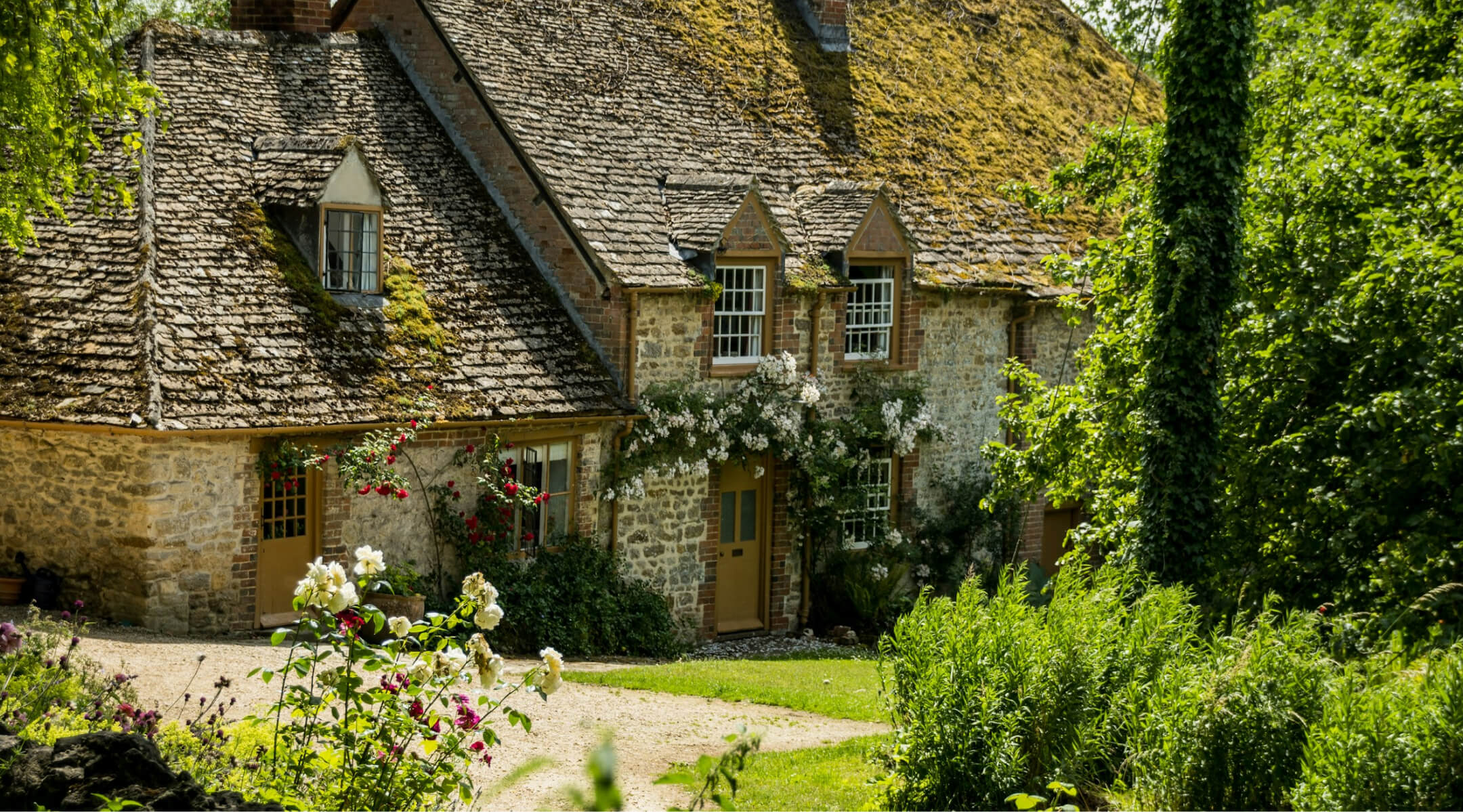 A classic, honey-colored limestone country home featuring weathered stone roof tiles, and white climbing roses framing the entrance.