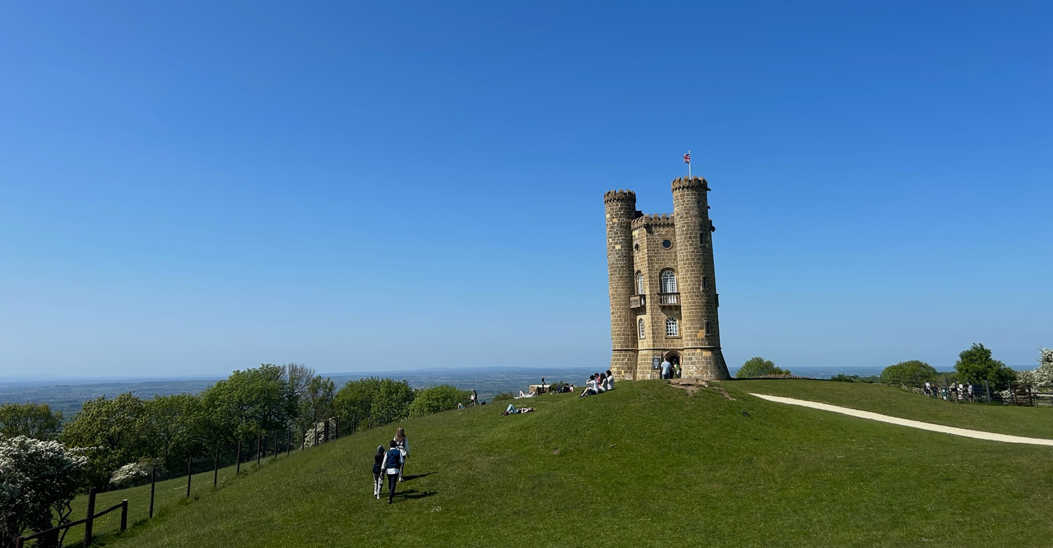 Broadway Tower on a grassy hill with people walking and sitting under a clear blue sky.