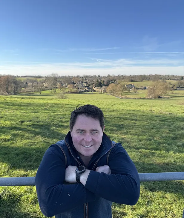 Ben, the founder of Corf Travel leaning on a farm gate on Leafield Farm with the village of Quenington behind him
