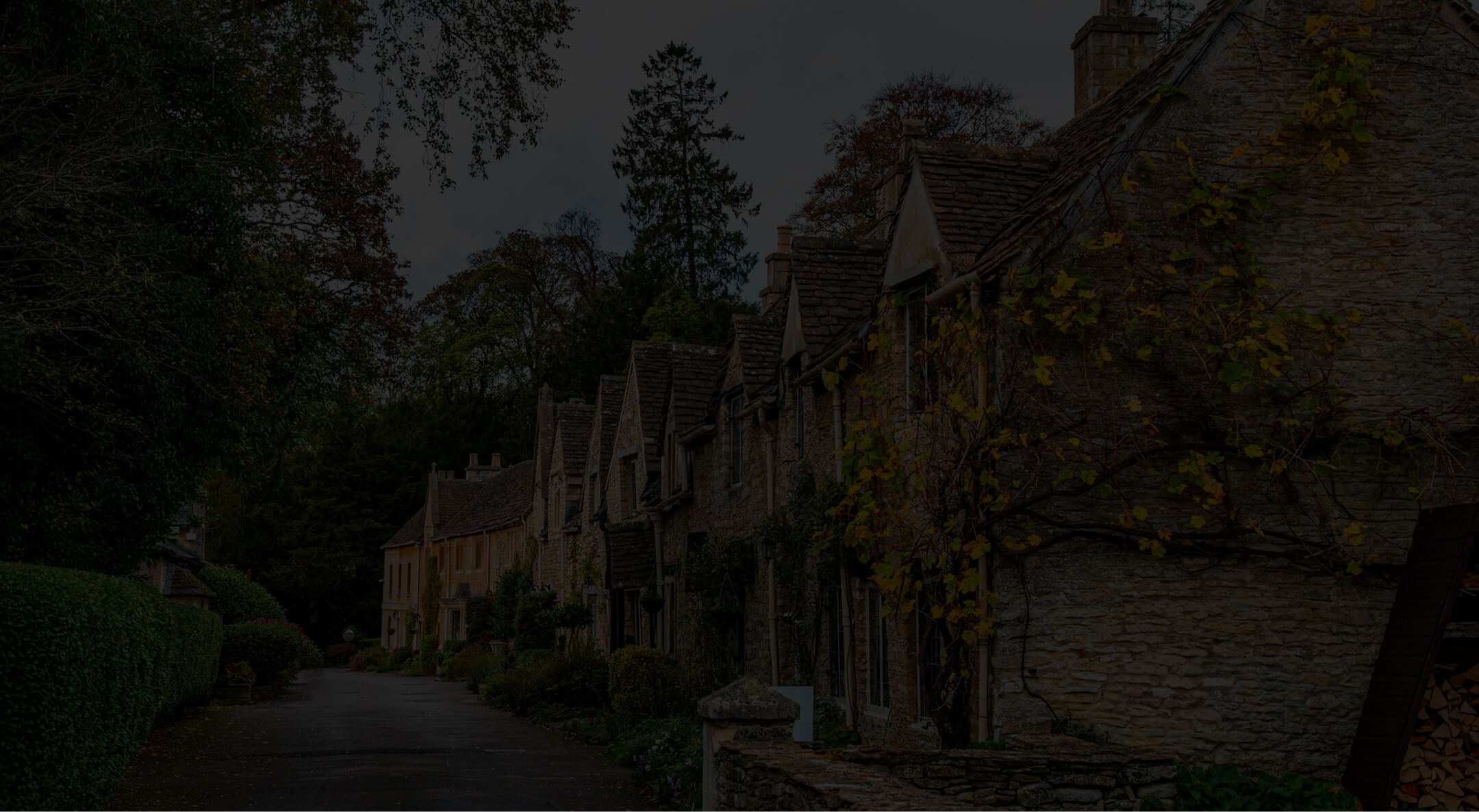 Stone cottages with pitched roofs line a quiet street bordered by hedges and trees in autumn.