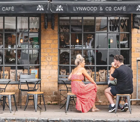 Two people sitting outside Lynwood & Co Café, a brick building with large windows and metal chairs.