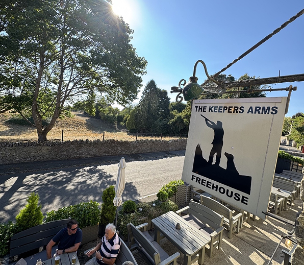 Sign for The Keepers Arms Freehouse with a silhouette of a man and dogs above an outdoor seating area.
