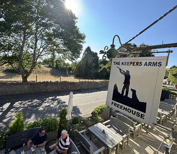 Sign for The Keepers Arms Freehouse with a silhouette of a man and dogs above an outdoor seating area.