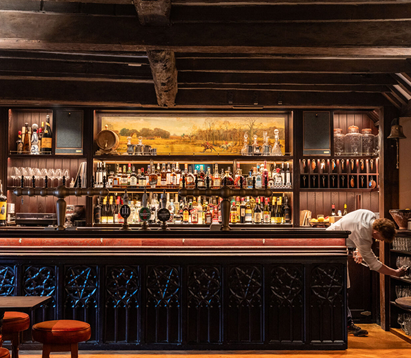 A classic pub bar with taps, bottles on shelves, a painting above, and a bartender reaching for a glass.