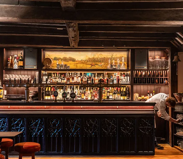 A classic pub bar with taps, bottles on shelves, a painting above, and a bartender reaching for a glass.