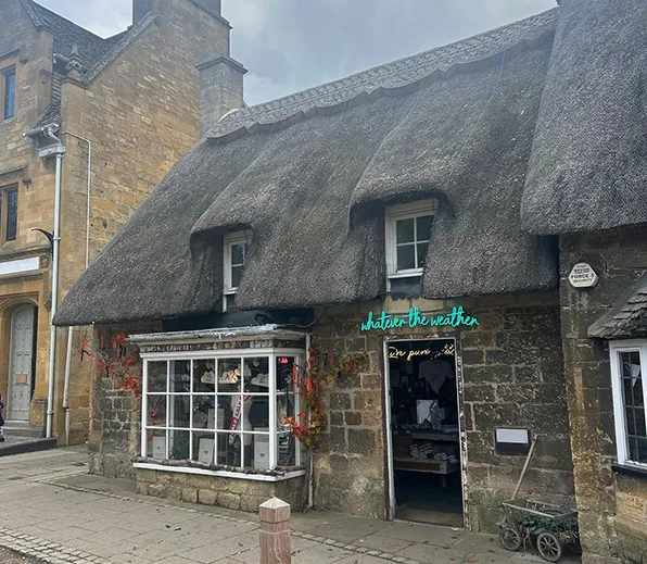 Stone building with thatched roof and 'Whatever the Weather' shop sign on a cloudy day.