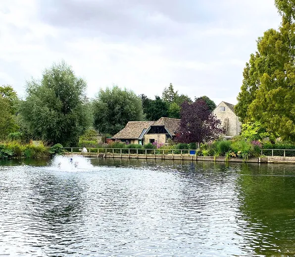 Waterfront view of a Bibury Trout Farm with trees and a fountain in the pond under cloudy sky.