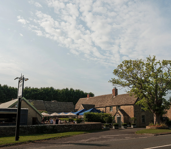 A countryside pub with outdoor seating under umbrellas beside a large tree on a partly cloudy day.