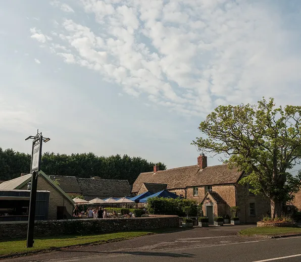 A countryside pub with outdoor seating under umbrellas beside a large tree on a partly cloudy day.
