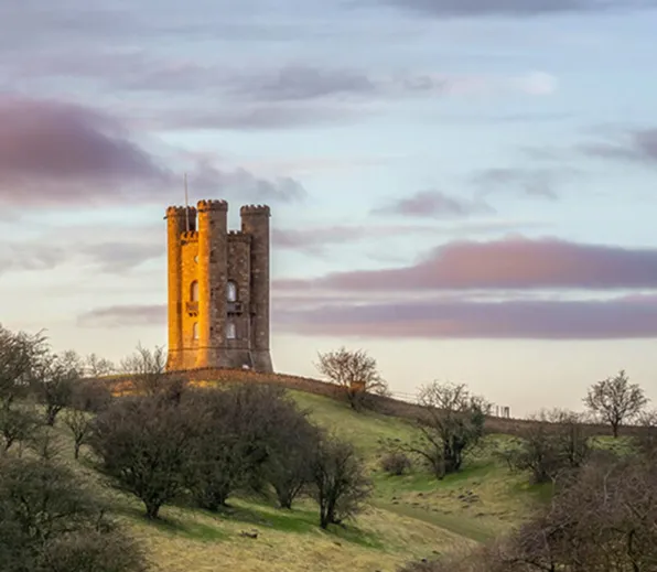 Broadway Tower on a grassy hill with trees under a cloudy sky during sunset.