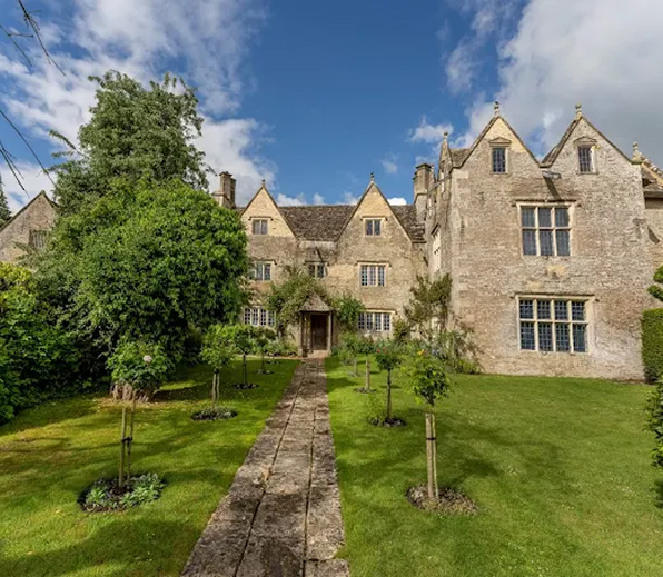 Stone manor house with steep gables, windows, and a walkway through a green garden under blue sky.