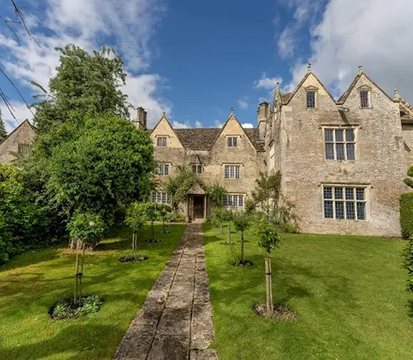 Stone manor house with steep gables, windows, and a walkway through a green garden under blue sky.