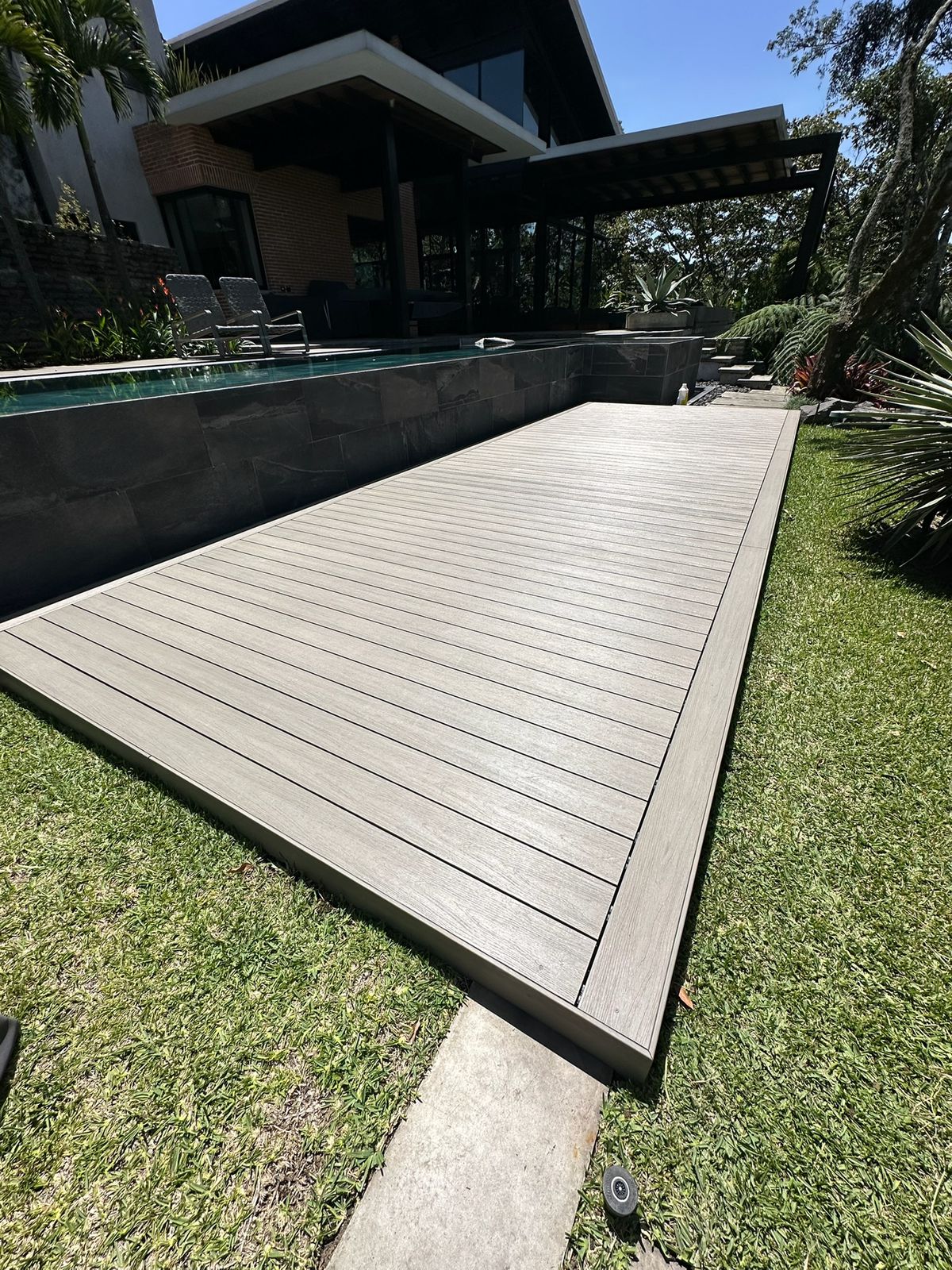 Piscina elevada con revestimiento oscuro y una terraza de madera rodeada de césped verde.