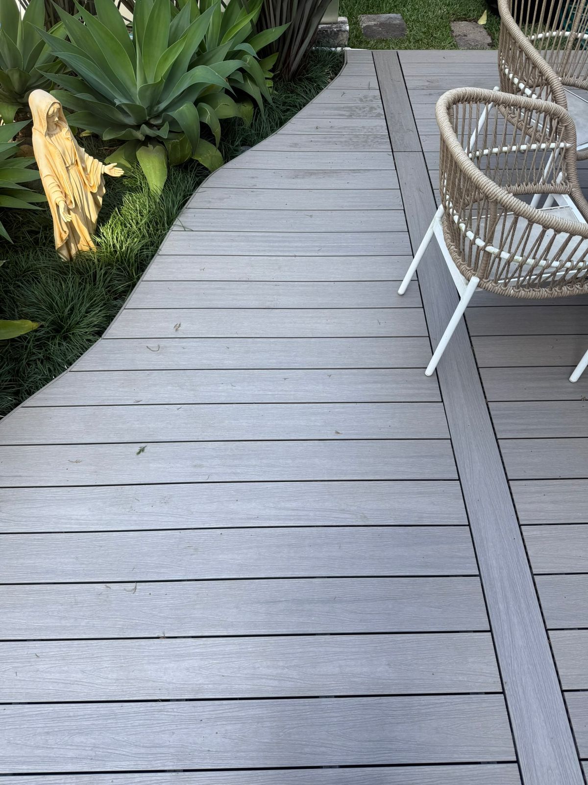 Piso de madera con dos sillas tejidas y una estatua religiosa amarilla entre plantas verdes.